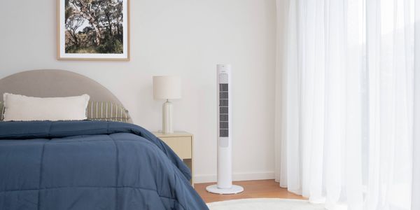 White tower fan standing beside a bed in a bright bedroom with sheer curtains, a bedside table, and a framed wall print