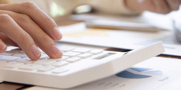 Close-up of a hand pressing buttons on a white calculator with financial charts and a laptop on a wooden desk.