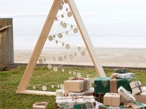 A wooden Christmas tree with decoration strung across it, presents wrapped in white, green and brown paper lie in front of the tree.