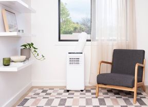Portable air conditioner positioned beneath a window in a bright living room, with an exhaust hose vented outside. A dark grey upholstered armchair sits beside the unit, while floating shelves with a potted plant and decorative items line the wall. A patterned rug covers the floor and sheer curtains frame the window.