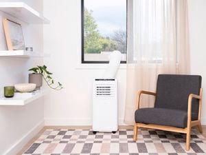 Portable air conditioner positioned beneath a window in a bright living room, with an exhaust hose vented outside. A dark grey upholstered armchair sits beside the unit, while floating shelves with a potted plant and decorative items line the wall. A patterned rug covers the floor and sheer curtains frame the window.