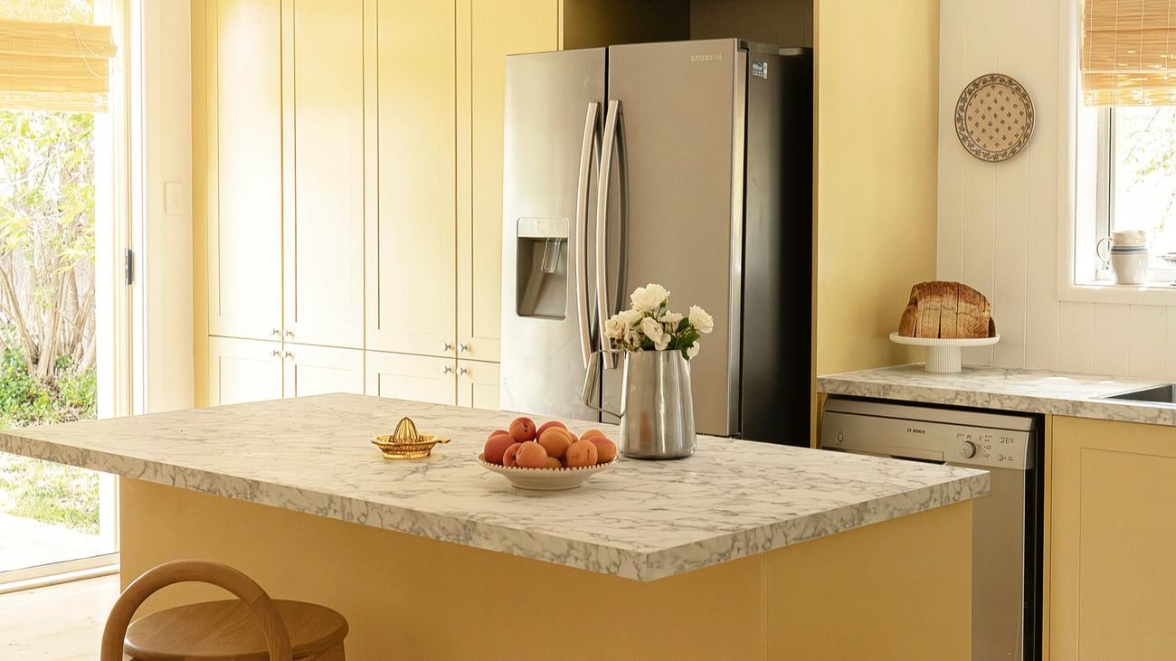 Alt text: A yellow kitchen with tall cabinets and a stainless steel double-door refrigerator. A marble island holds a bowl of peaches and a vase of white flowers, with a wooden stool underneath and a dishwasher and loaf of bread on the side counter.