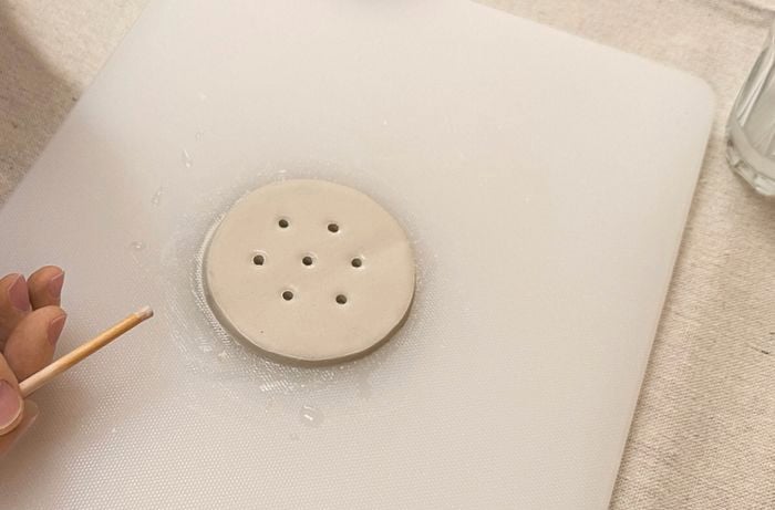 A hand holding a toothpick next to a circular piece of clay with holes poked into it.