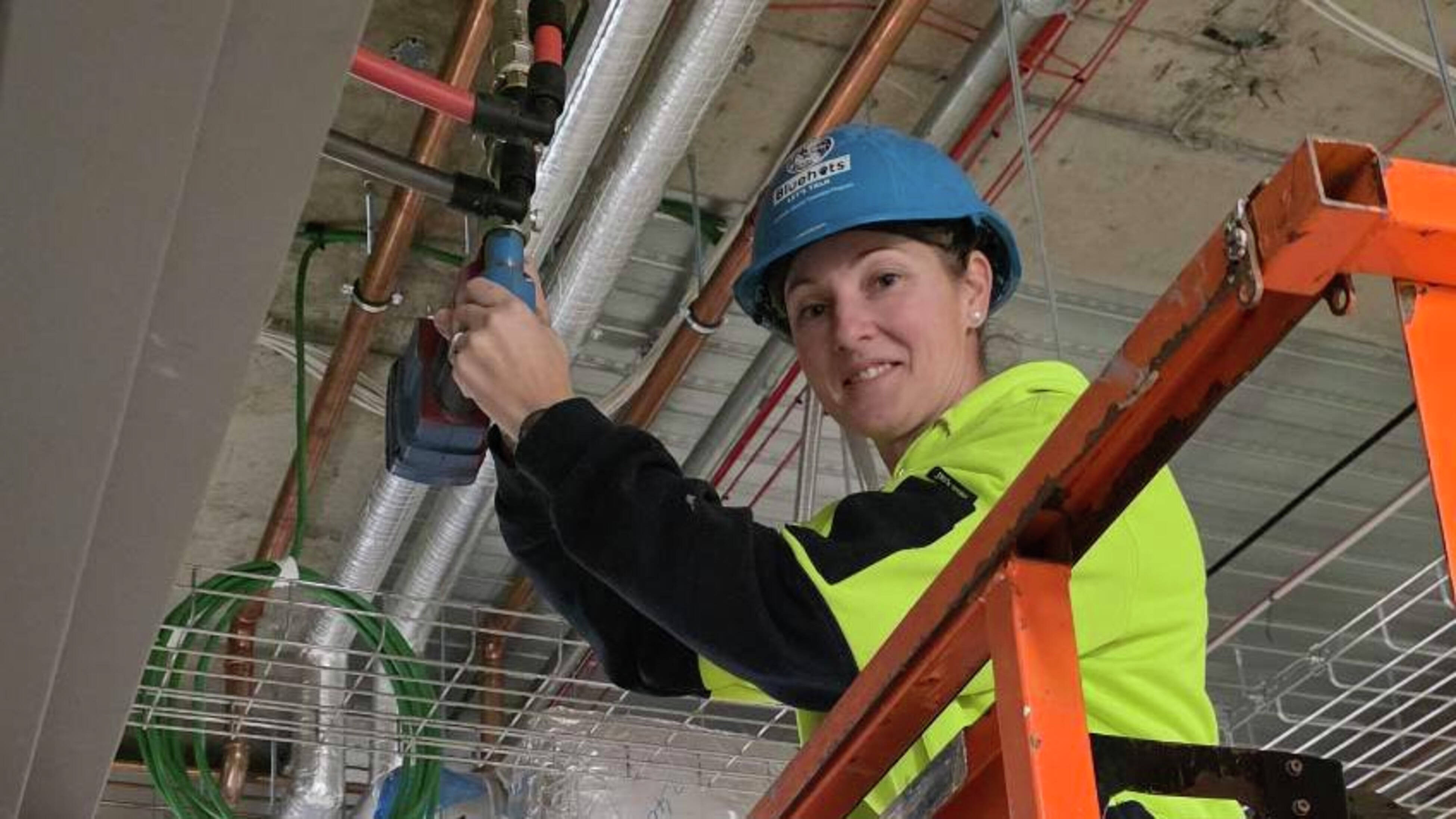 A female plumber wearing yellow hi-vis and a blue helmet while fitting REHAU pipes.