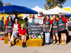 Group of Bunnings Team Members standing and sitting around a Bunnings Community Grants sign at an outdoor community event with market tents in the background.