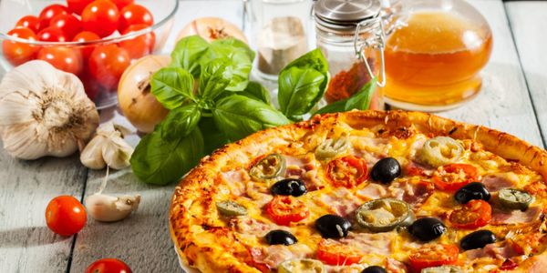 Freshly baked pizza topped with cherry tomatoes, olives and jalapeños, surrounded by garlic, basil, onions and other fresh ingredients on a rustic wooden surface.