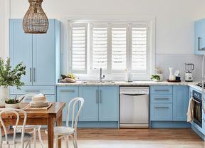 A coastal kitchen with blue cabinets, white shutter blinds, and a wooden table with chairs.