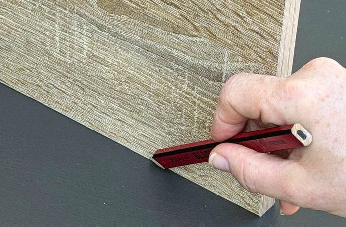  A close up of a hand, holding a pencil and making a mark alongside a piece of plywood.