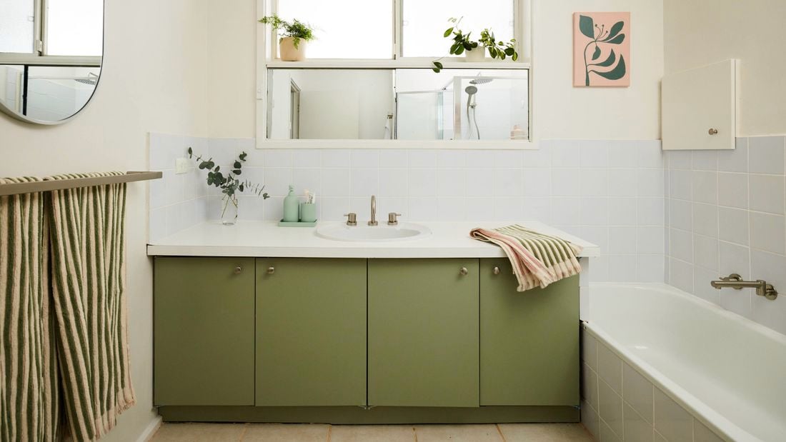 Bright bathroom with a green vanity cabinet and white benchtop, featuring a central sink beneath a wide mirror, a bathtub to the side, white tiled walls, striped towel on a rack, and potted plants along the windowsill for natural light and greenery.