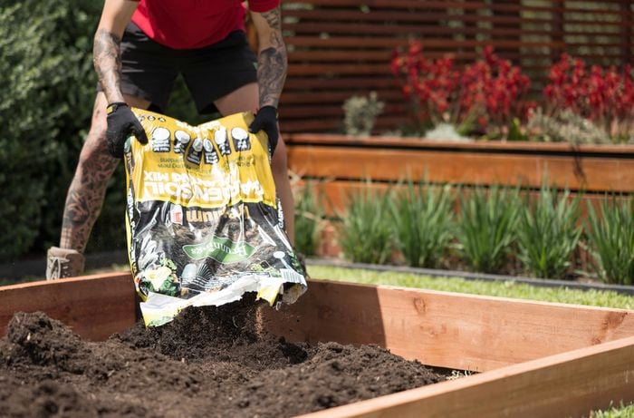 A person wearing a red shirt and work boots is filling a bag of premium garden soil mix into a raised wooden garden bed. 