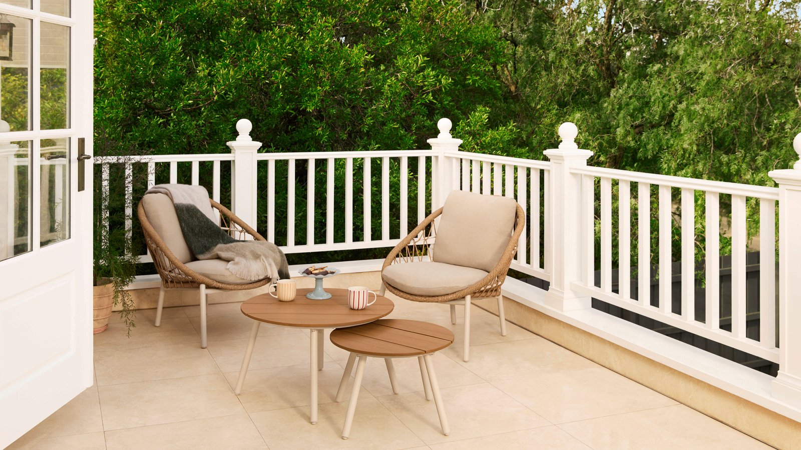 Outdoor deck with white balustrade, two woven lounge chairs, a round coffee table with mugs, and leafy garden views.