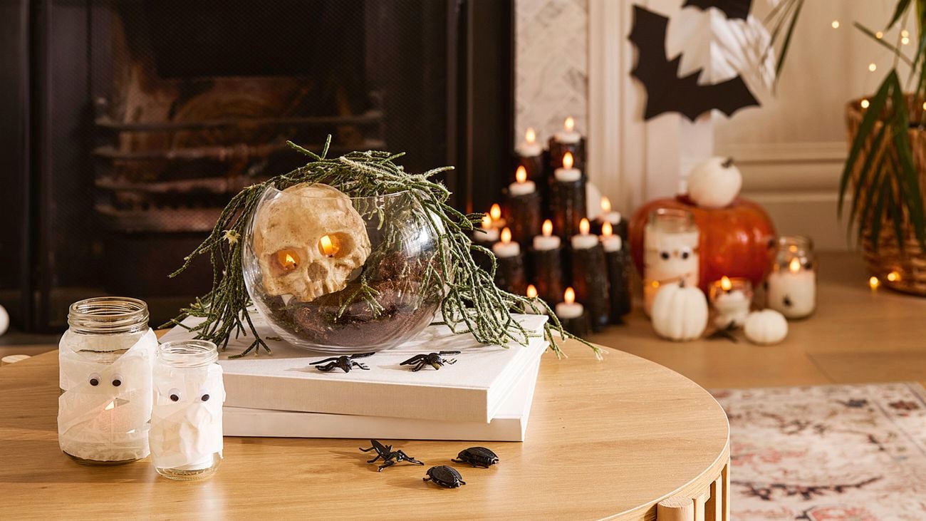 A wooden table with Halloween decorations, including jars wrapped like mummies, a skull in a glass bowl, plastic insects and books. Candles and pumpkins are visible near the fireplace in the background