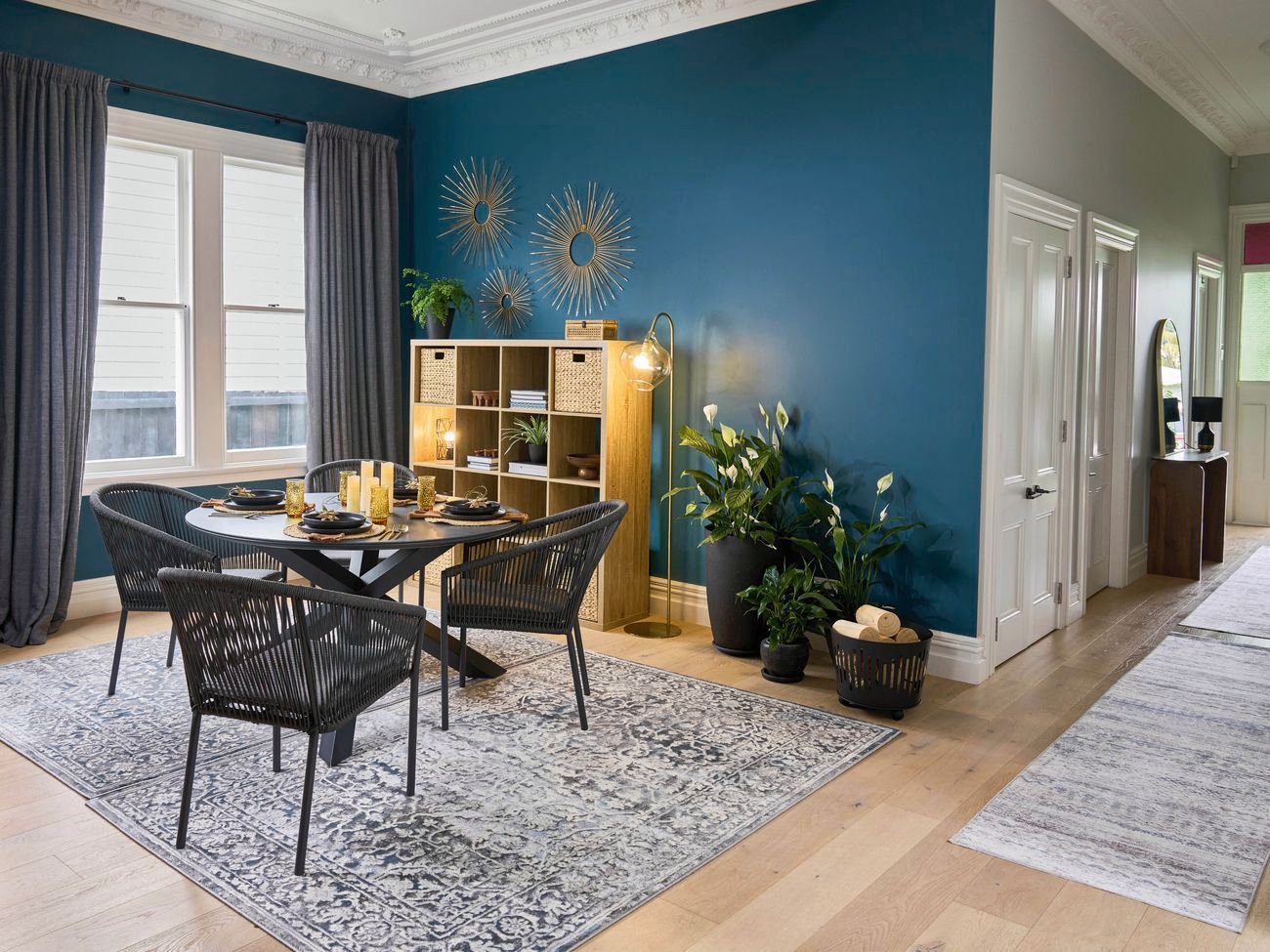 Modern dining room with deep teal feature wall, round table set with black tableware, woven chairs, shelving unit, indoor plants, and decorative gold wall art.