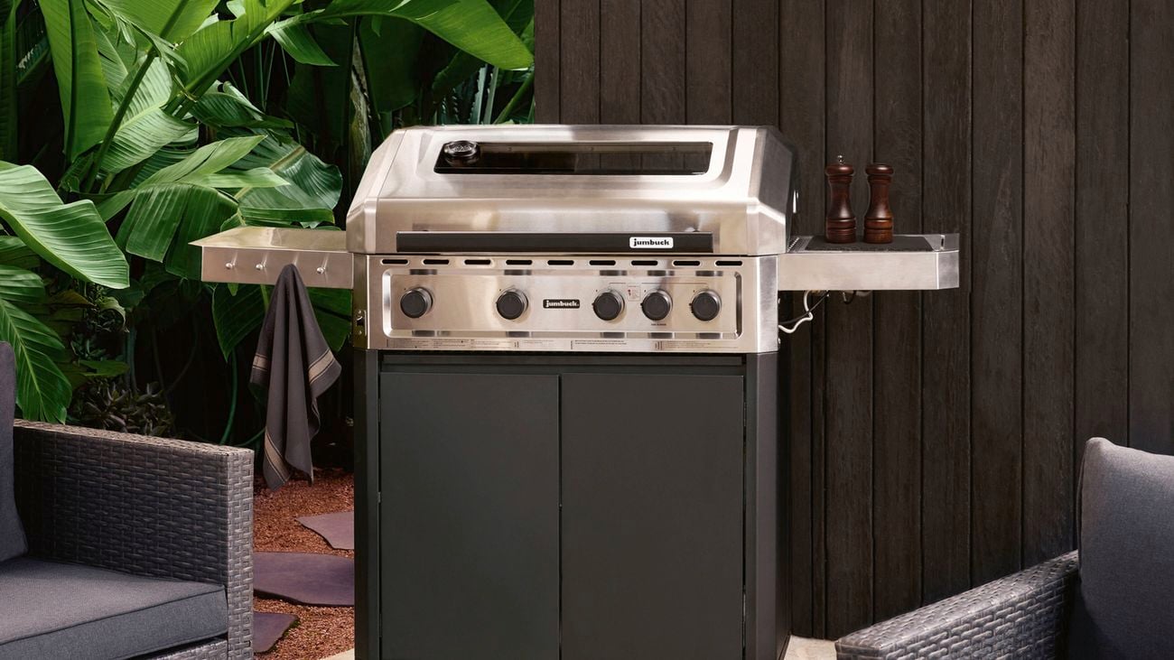 Stainless steel barbecue grill on a paved patio, surrounded by lush green foliage and a dark timber wall