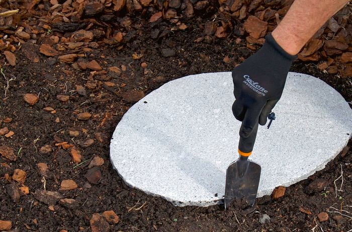 A person wearing gloves uses a small trowel to dig soil around a circular concrete slab in a garden. 