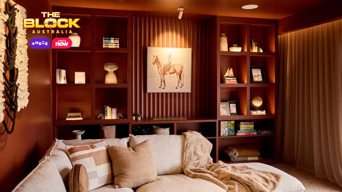 A cozy living room with a sectional sofa decorated with pillows and a blanket. Behind the sofa, built-in shelves display books, artwork, and decorative items, with a prominent horse painting at the center. The room features warm lighting and earthy tones, creating a welcoming ambiance.