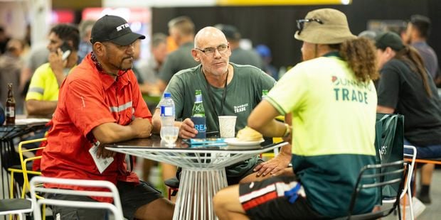   A group of people sit at a table at the trade expo, talking and having food and drinks.