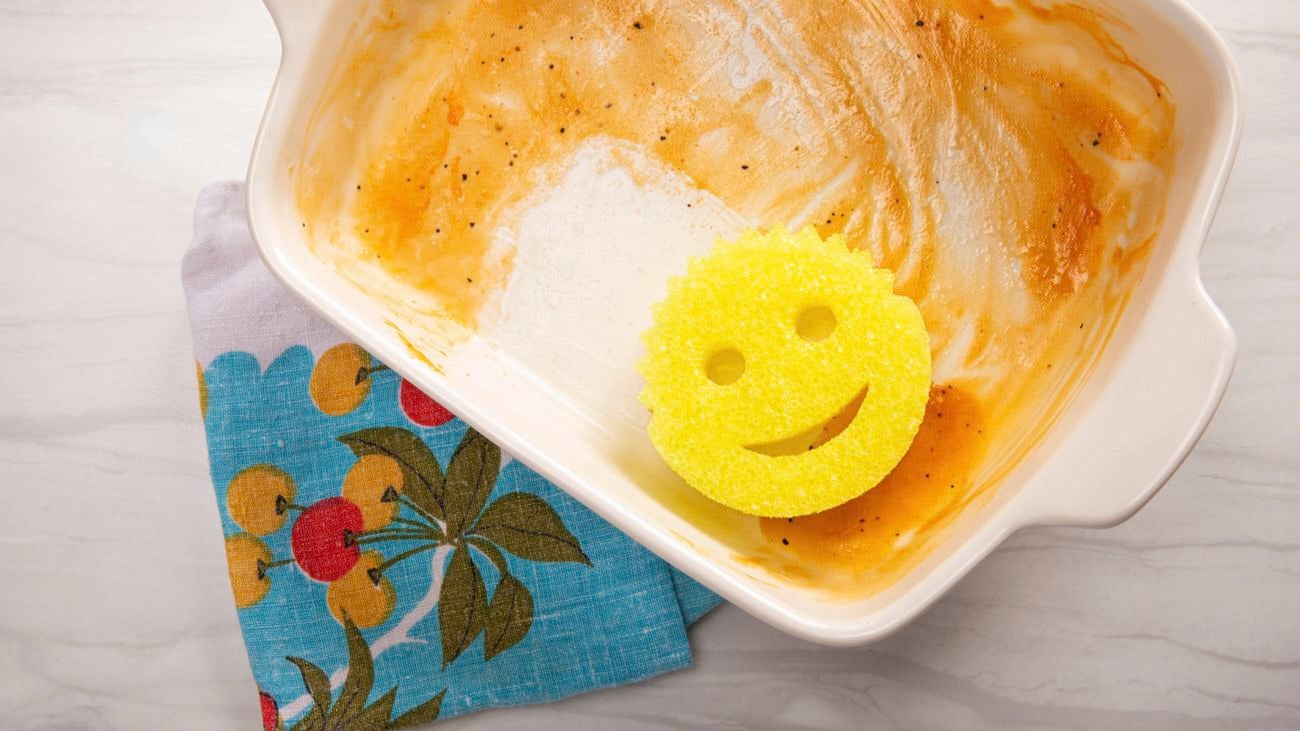 A yellow Scrub Daddy sponge cleaning a baking dish with a colourful towel underneath.