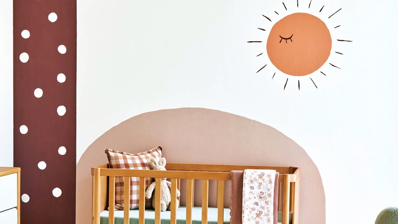 White walled bedroom with a wooden cot, dresser and green rocking chair. The walls are painted with patterns of a dusty colour palette and toys are strewn on the floor.