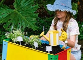 A child wearing a blue sunhat and gardening gloves kneels in a garden, planting seedlings in a brightly coloured raised planter box and watering them with a small yellow watering can