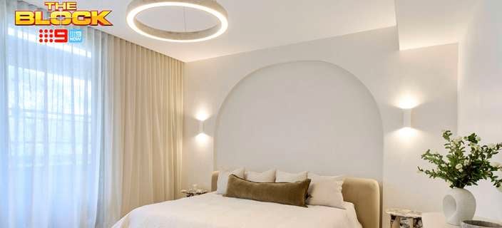 A furnished bedroom with a bed, lights on the wall and ceiling, side table and another table with a vase on it with flowers in it.