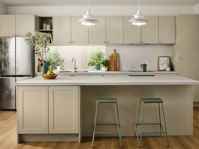 Beige kitchen with a white marble benchtop, two green stools at the bench, with multiple vases with flowers in them on the benchtop.