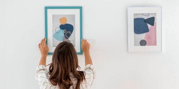 Person hanging a framed abstract artwork on a white wall next to another framed piece, showcasing a minimalist interior design.