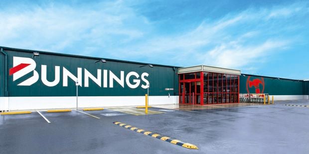 Exterior view of a Bunnings Warehouse store with large white “BUNNINGS” signage on a green facade, a red-framed glass entrance, and a mostly empty car park in the foreground under a blue sky with light clouds.