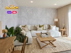 A modern living room with a large white sectional sofa decorated with neutral and green pillows. A wooden coffee table with a decorative tray sits in front, and a side table with a lamp and vase is placed to the right. Light-colored curtains cover large windows, and palm tree wallpaper adds texture to the walls. A desk with drawers and a chair is positioned on the left, near potted plants. 