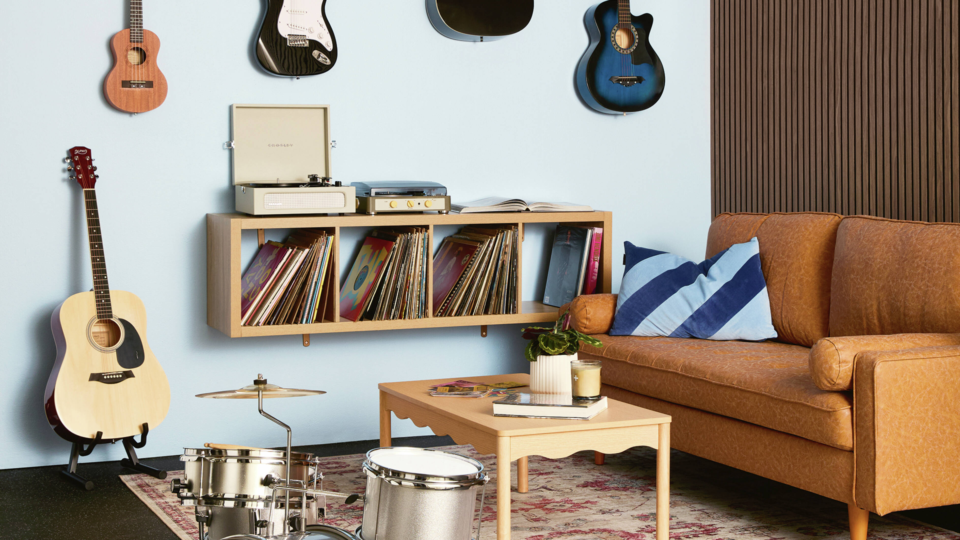 A music room with acoustic wall panelling, rubber floor matting, a tan sofa, drum kit, record player on floating shelves and wall-mounted guitars