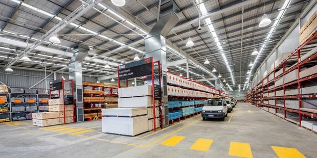 Wide aisle inside a Bunnings warehouse with high shelving stocked with building materials, marked traffic lanes, and utility vehicles parked along the centre aisle.