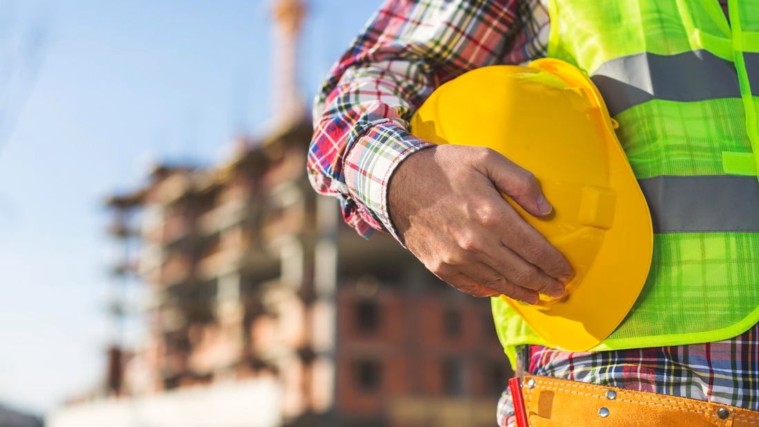 Close up shot of the torso of a construction worker in high vis vest holding a hard hat, in front of a building site