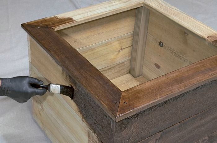 Hands holding a paintbrush to stain a wooden planter box a dark brown colour.