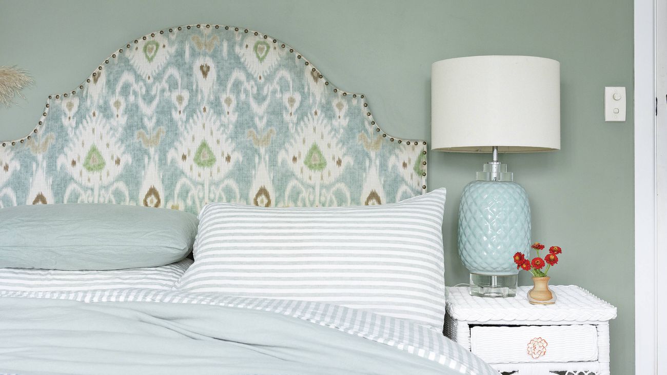 Bedroom with patterned headboard, striped pillow, light blue bedding and blue lamp on a white bedside table.