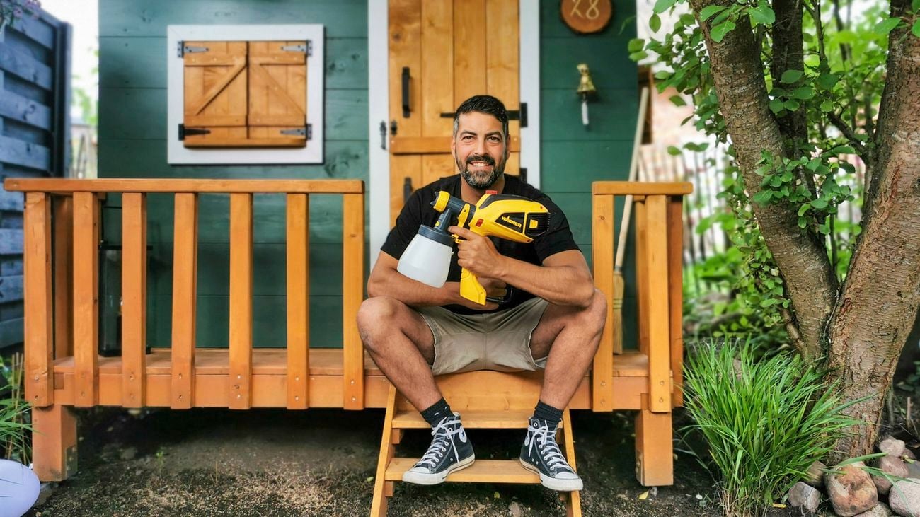 A man in a black t-shirt and tan shorts holding a Wagner paint sprayer sits on the steps of a garden cabin