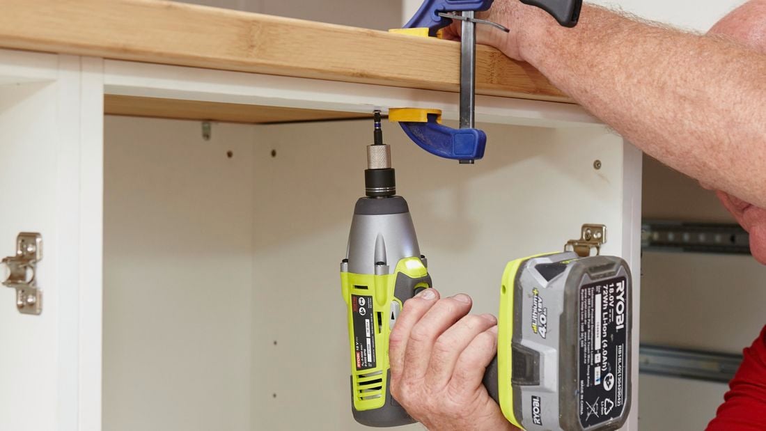 A drill being used to create screw holes to anchor down a bamboo benchtop, currently held in place with a clamp