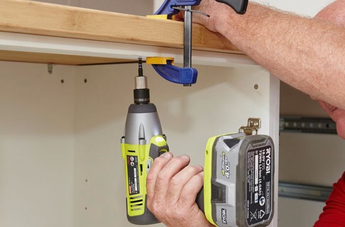 A drill being used to create screw holes to anchor down a bamboo benchtop, currently held in place with a clamp