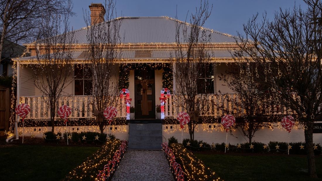 Cream traditional Australian style home with warm fairy lights across the roof, veranda and bushes along with light up candy canes as well as red and white decorative lollipops.