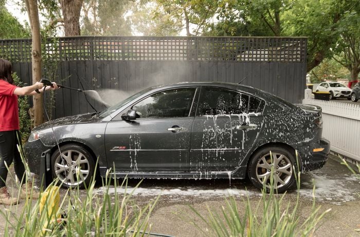 A woman using a pressure washer to rinse off the soap from the car.]