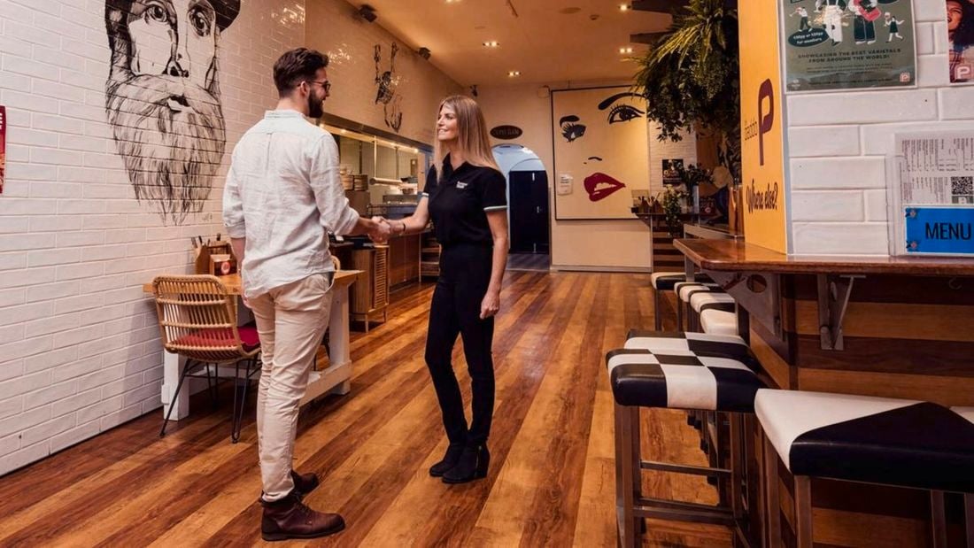 Two people shaking hands inside a modern café or restaurant with wooden floors, wall murals, bar seating, and posters visible along the walls.