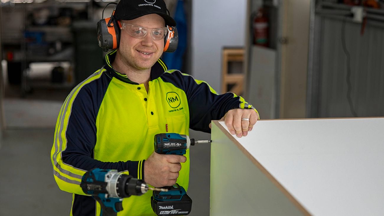 A man wearing a hi-vis long-sleeved top, safety glasses, ear protection and a baseball cap smiles at the camera while holding a Makita drill up to the side of a white cabinet