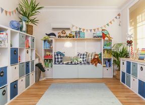 Bright, organized playroom with blue-and-white storage cubbies, toys on shelves, a light rug, and large windows letting in natural light.