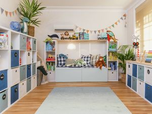 Bright, organized playroom with blue-and-white storage cubbies, toys on shelves, a light rug, and large windows letting in natural light.