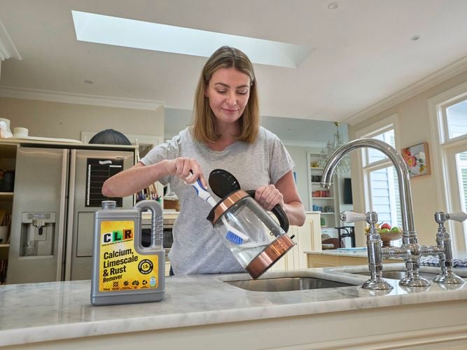 A lady holding a cleaning brush in a kettle with a bottle of CLR Calcium Limescale & Rust 1L on the kitchen bench.