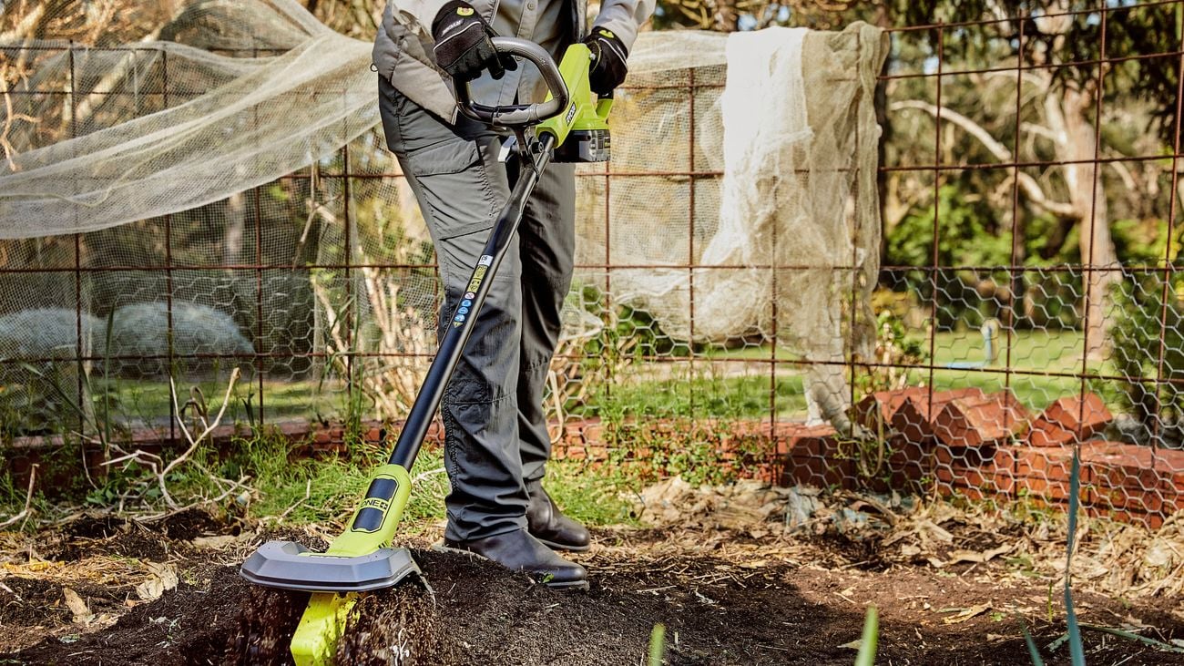 A person holding a cultivator on top of soil in a backyard.