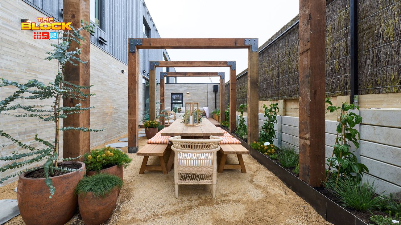 Outdoor dining area featuring a long timber table and benches beneath a series of timber pergolas, surrounded by raised garden beds and potted plants.