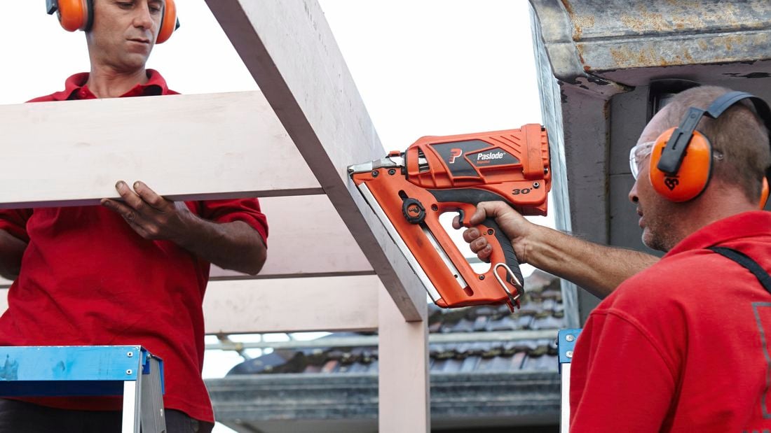 One person on a ladder holding a rafter while another person attaches it to a beam using a nail gun