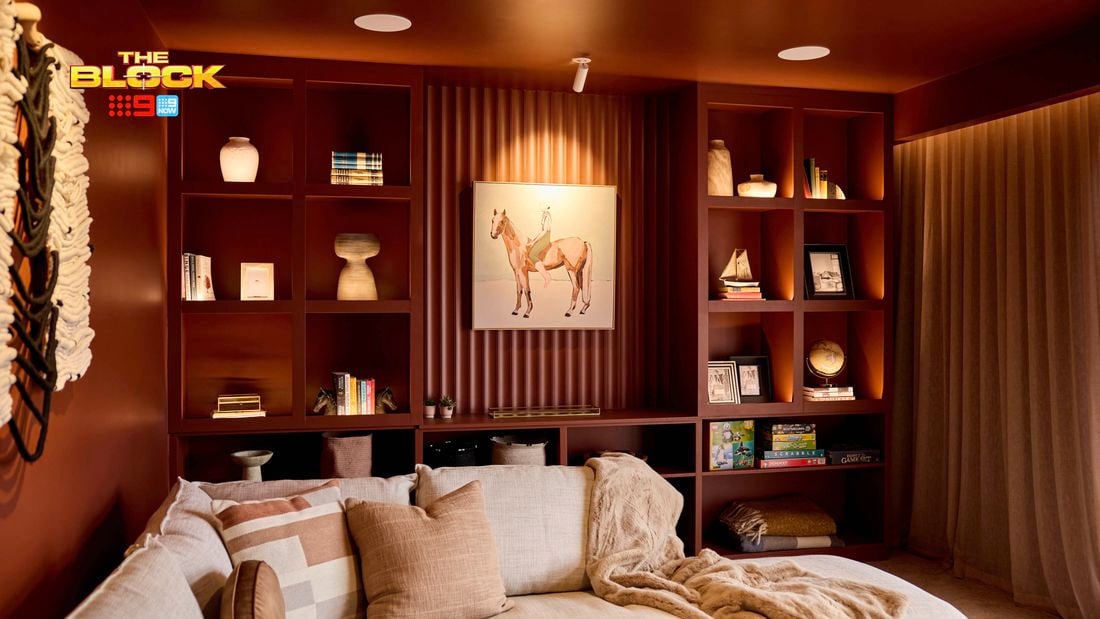 A cozy living room with a sectional sofa decorated with pillows and a blanket. Behind the sofa, built-in shelves display books, artwork, and decorative items, with a prominent horse painting at the center. The room features warm lighting and earthy tones, creating a welcoming ambiance.