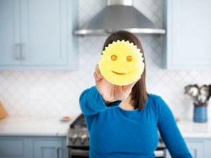 A woman holding a yellow Scrub Daddy sponge in front of her face, with a blue kitchen in the background.