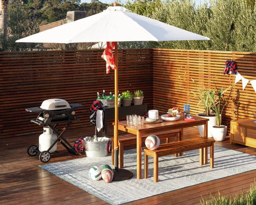 Outdoor entertaining area on a timber deck featuring a large white market umbrella, wooden dining table with bench seating, portable gas barbecue, drinks in ice buckets and decorative plants. The space is styled for backyard entertaining with sporting balls, outdoor cushions and a timber privacy screen surrounded by trees.